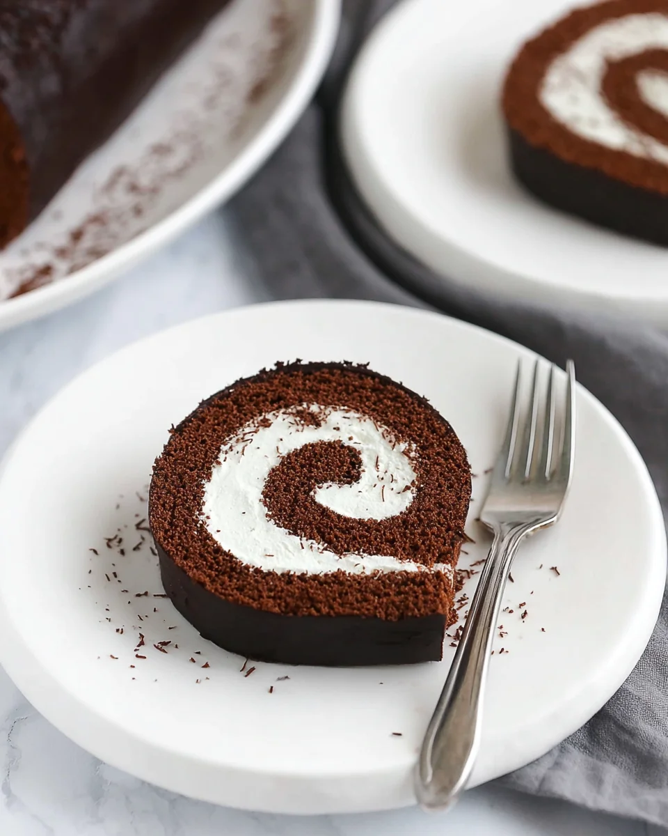 With a fluffy cake, creamy filling, and rich chocolate ganache, this Chocolate Swiss Roll recipe has all the elements of the classic treat. A slice of chocolate Swiss roll on a white plate