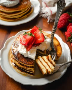 stack of cottage cheese pancakes topped with whip cream and fresh strawberries, with a fork full cut out.
