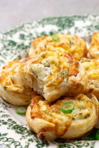 A close-up of golden-brown pastry swirls filled with a cheesy herb mixture, garnished with sliced green onions. The pastries are on a white and green patterned plate.
