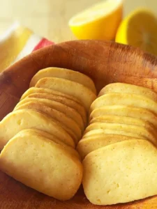 Lemon slice-and-bake cookies in a row on a wooden bowl, halved lemons in the background.