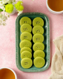 A rectangular green tray with 2 rows of green matcha shortbread cookies. There are 2 cups of tea and a bouquet of flowers nearby.