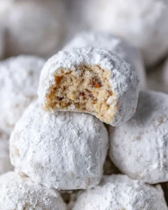 Pecan snowball cookies piled on a plate. The cookie on top has a piece taken out so you can see the light brown interior and pieces of pecans