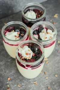 Four mini cups of high protein vanilla pudding topped with whipped cream, nuts, and mixed berries on a counter.