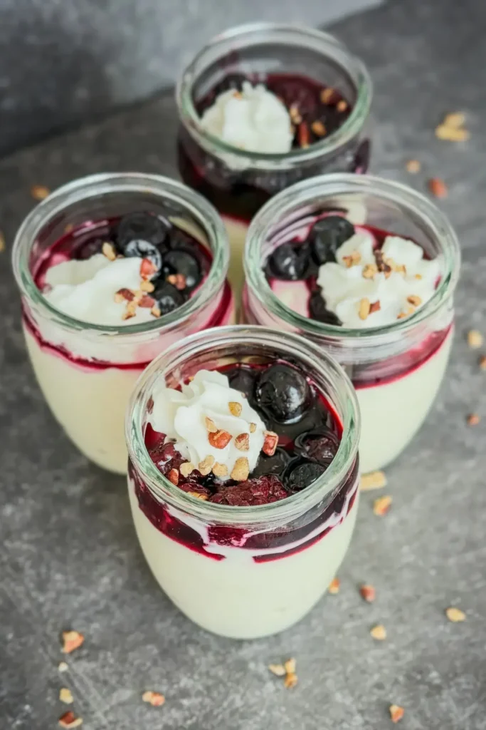 Four mini cups of high protein vanilla pudding topped with whipped cream, nuts, and mixed berries on a counter.