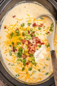 Hearty potato soup in a black slow cooker topped with cheese, green onion, bacon and black pepper with a ladle.