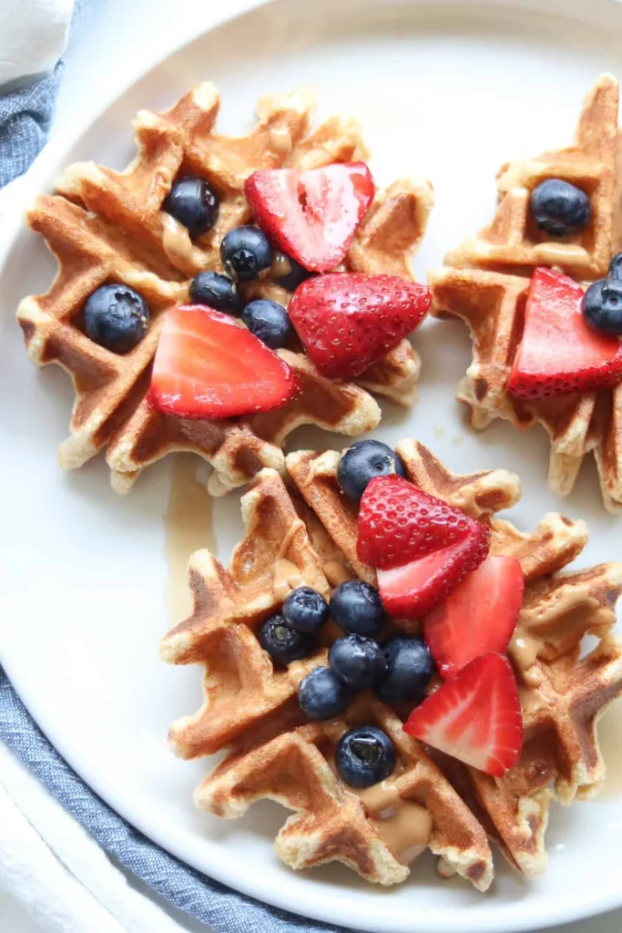 Cooked cottage cheese waffles on a white plate drizzled with peanut butter and topped with slices of fresh strawberries and blueberries.