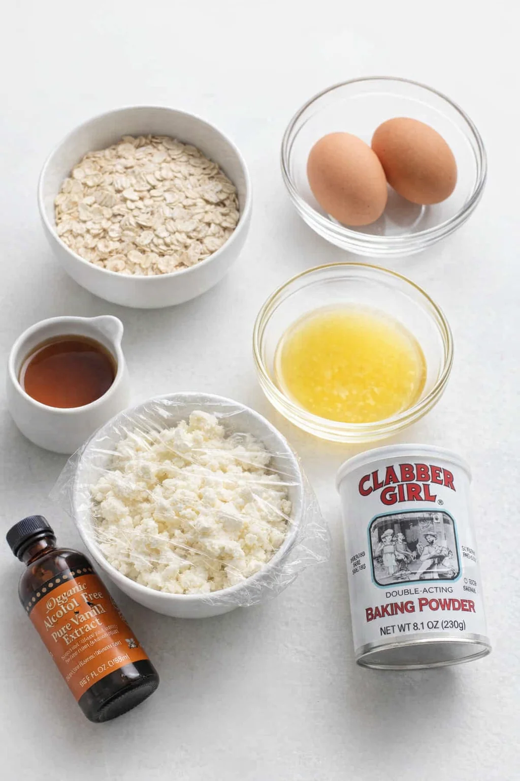 Arrangement of baking ingredients including oats, eggs, butter, cottage cheese, vanilla, baking powder, maple syrup, and flavoring on a white background.
