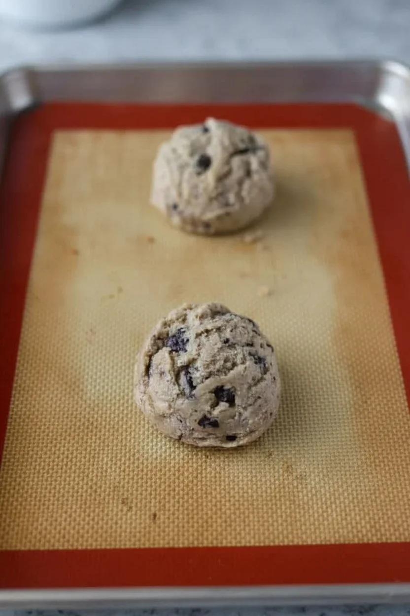 Two raw cookie dough balls with chocolate chips on a silicone baking mat with a red border.