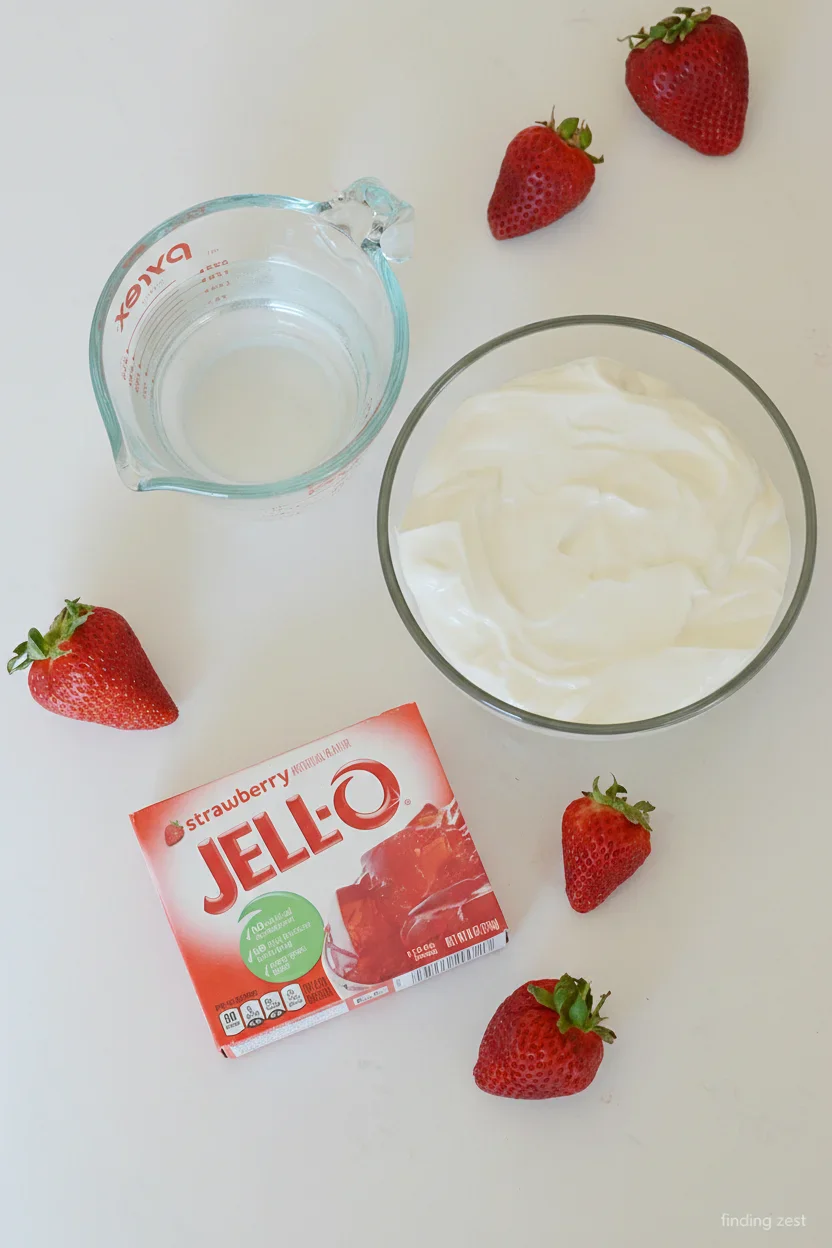 Fresh strawberries, a measuring cup with water, a bowl of yogurt, and a box of strawberry Jell-O on a white surface.