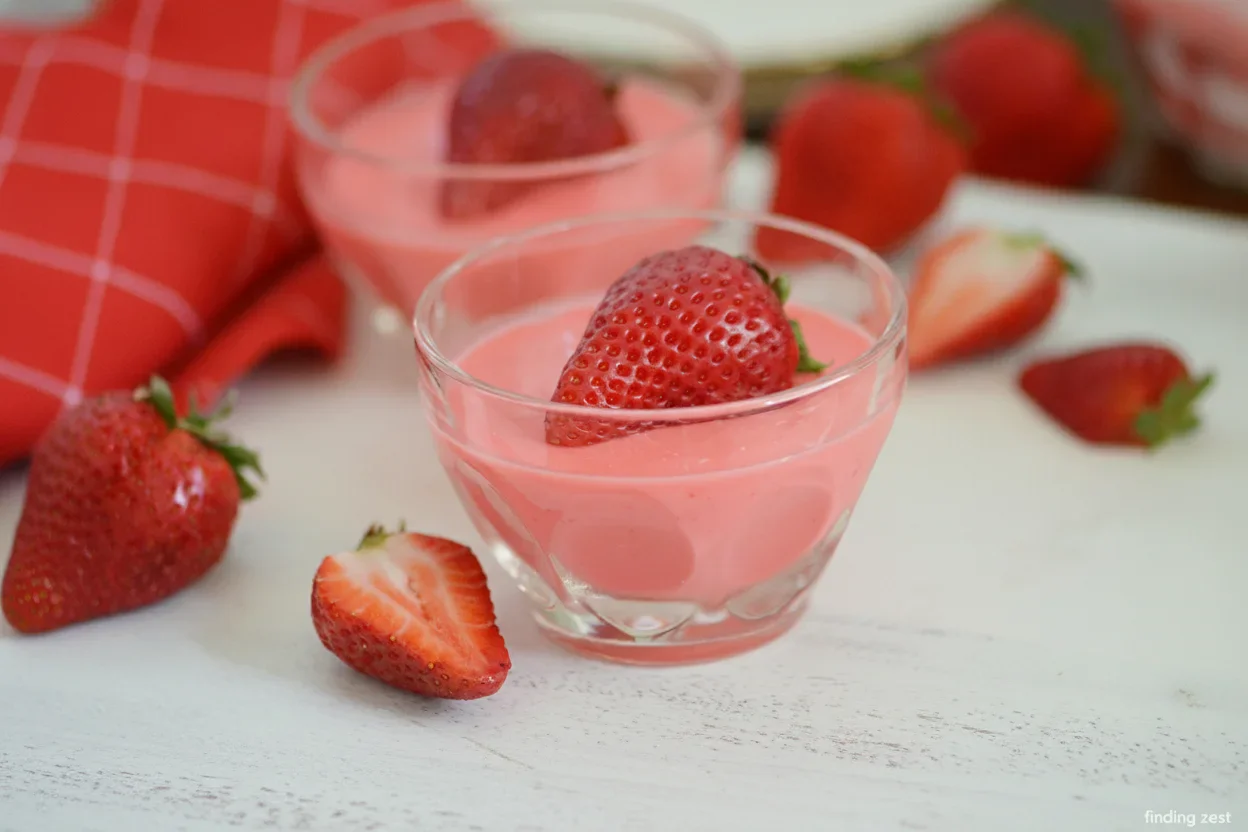 Two glass bowls of pink strawberry dessert topped with fresh strawberries, surrounded by scattered strawberries on a white surface.
