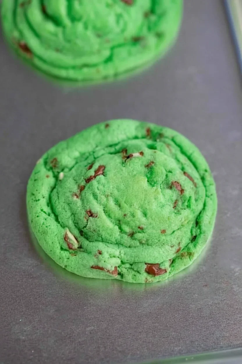 Bright green cookie with chocolate chunks on a gray baking sheet, with another cookie in the background.