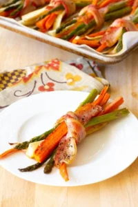 Roasted bacon wrapped vegetables on a white plate with a piece of bacon wrapped around it.