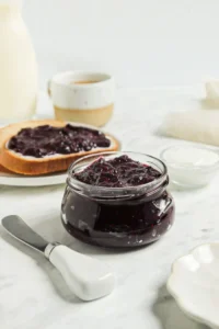 Homemade berry chia seed jam in a glass jar with bread and dairy products in the background.