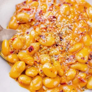 Close-up of tender butter beans in a thick, orange-colored sauce with visible herbs and chili flakes, served on a plate with a spoon.