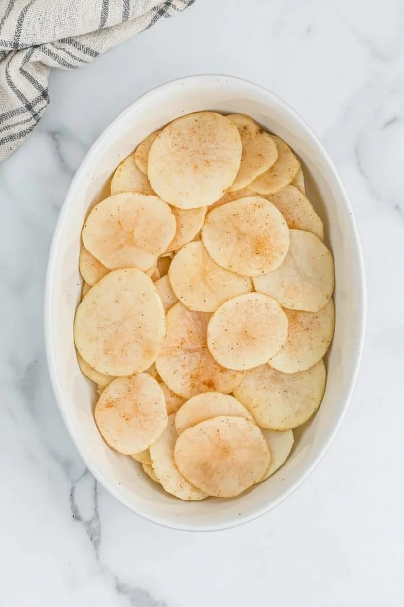 Thinly sliced potatoes sprinkled with paprika in a white oval dish on marble surface.