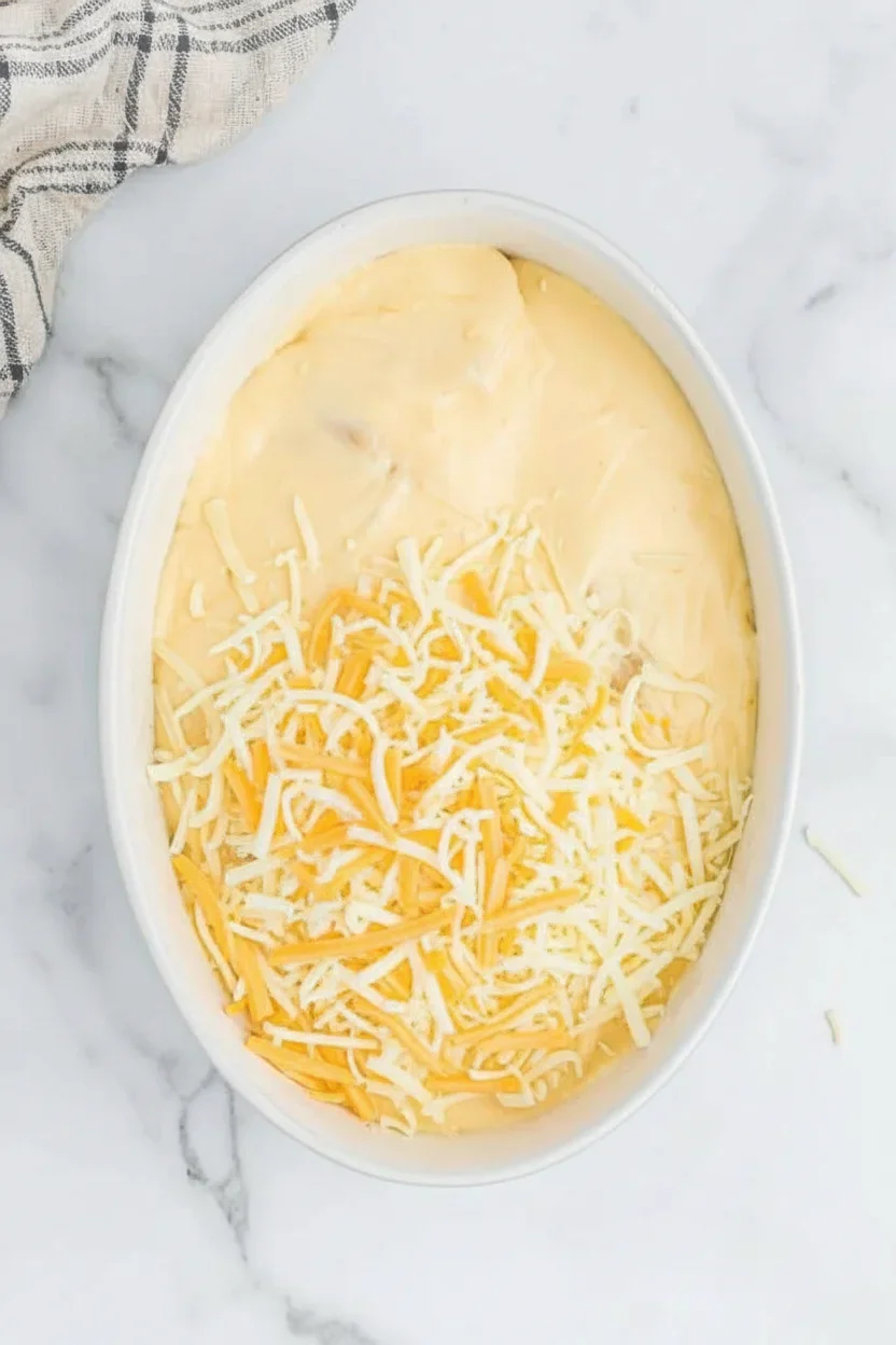 Oval dish filled with creamy cheese mixture topped with shredded cheddar and white cheese on a marble surface.