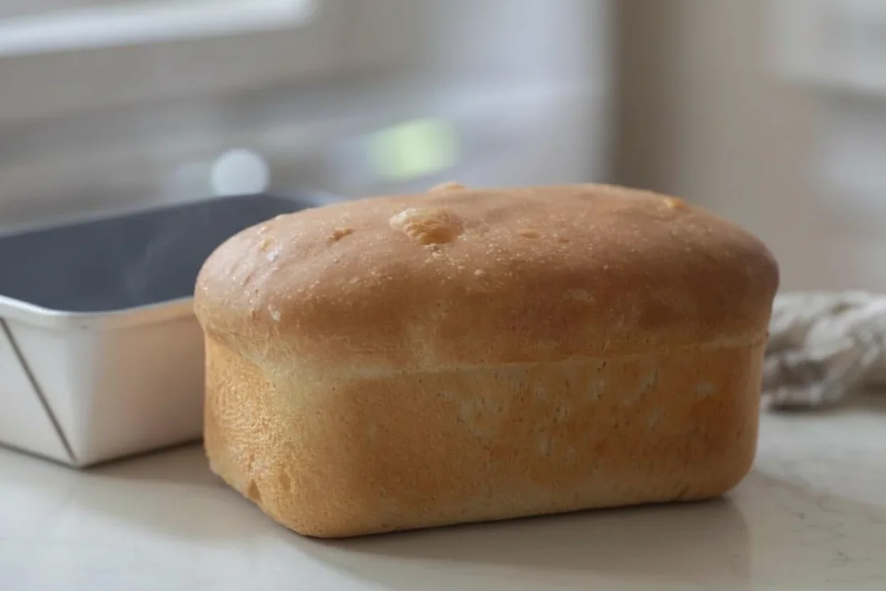 A golden-brown loaf of bread with a smooth crust on a white surface in a kitchen setting.