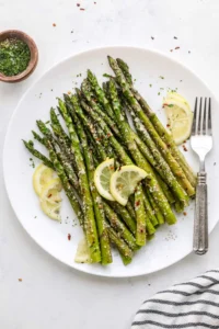 Roasted asparagus with parmesan piled up on a round white plate with sliced lemon in it and a silver fork next to it.