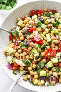 Mediterranean Chickpea Salad tossed in a large salad bowl with silver serving spoons in the bowl and a bowl of chopped parsley behind it.