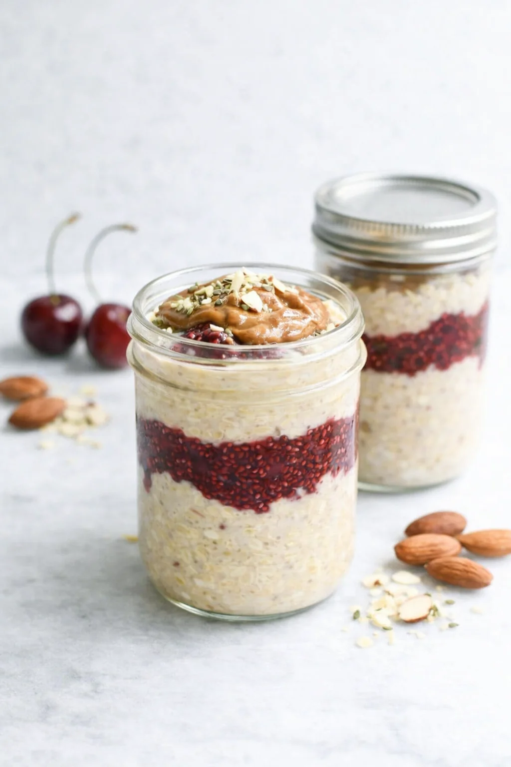 Two glass jars show layered overnight oats with a berry layer and a nut-butter topping, topped with sliced almonds.