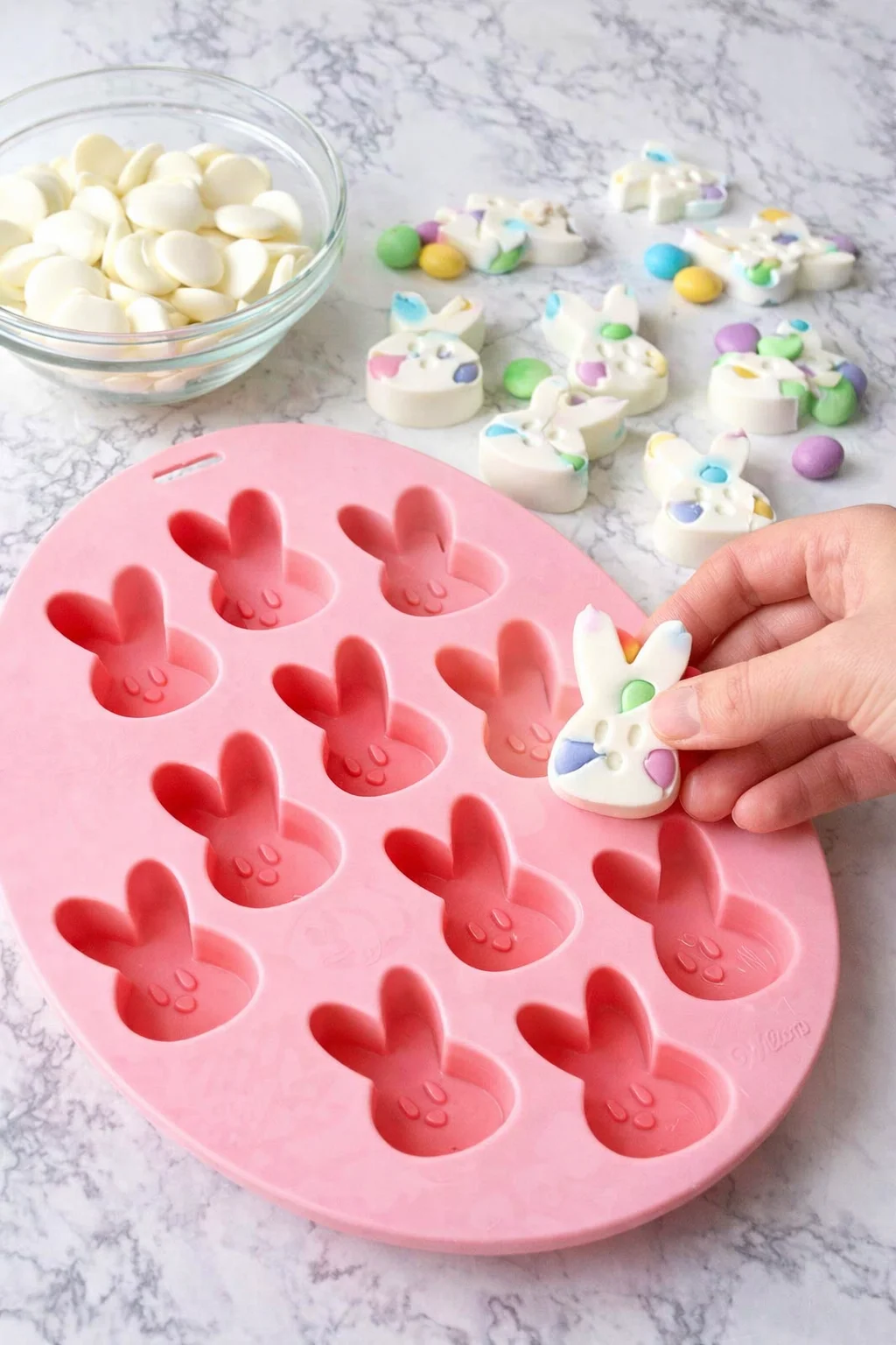 Hands flexing silicone mold as small white bunny candies pop out onto a baking sheet.