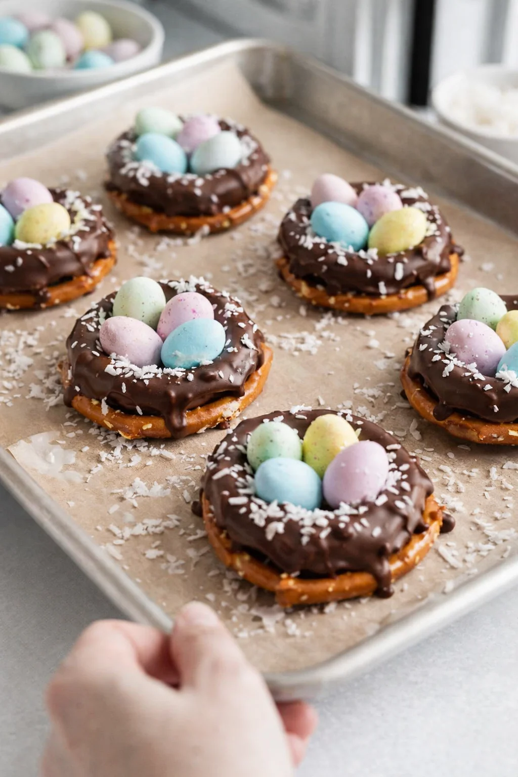 step-process-placeholder-5-6.png Mini candy eggs placed into pretzel nests, with coconut or sprinkles visible and baking sheet chilling.