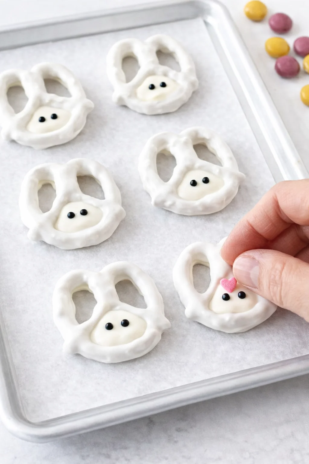 White-coated pretzels chilled on tray; piping bag fills bottom openings and sprinkles pressed for bunny faces.