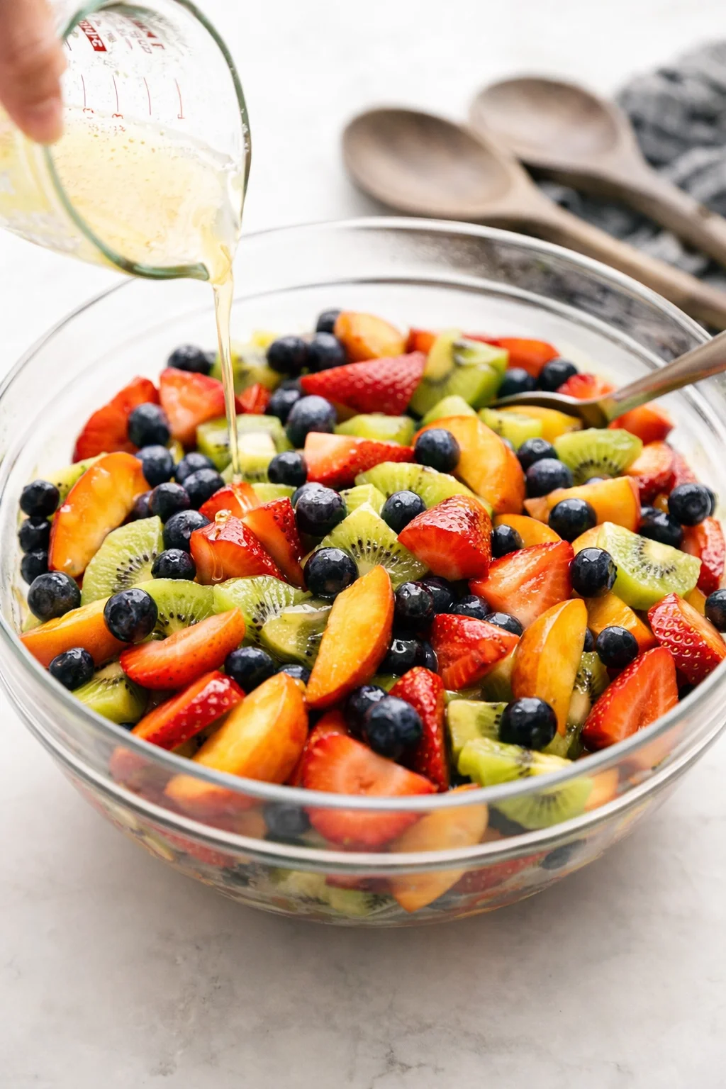 Hand drizzling honey-lime dressing over mixed fruit while a spoon readies to gently toss.
