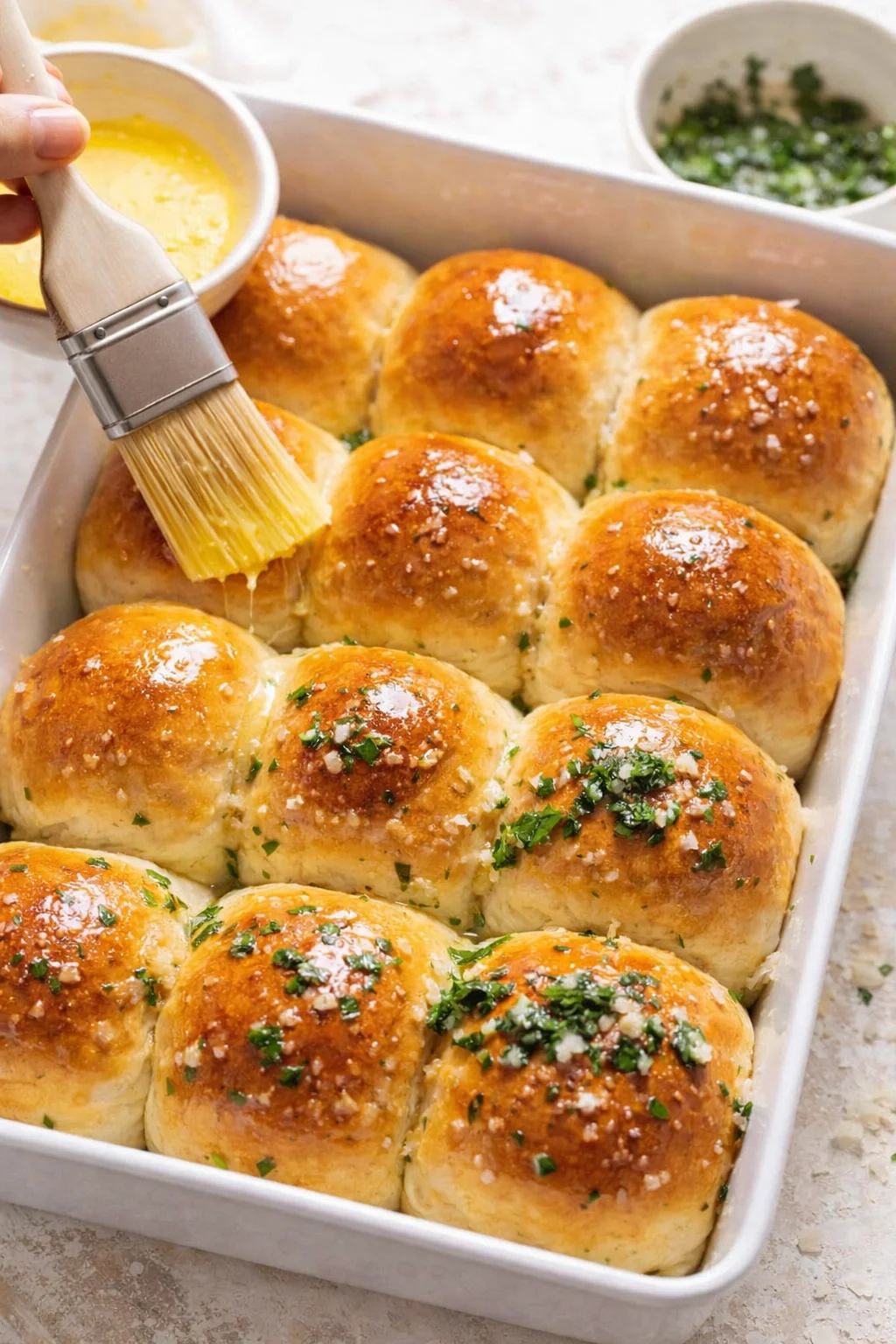 Freshly baked golden rolls being brushed with melted garlic-parsley butter, flaky salt visible.