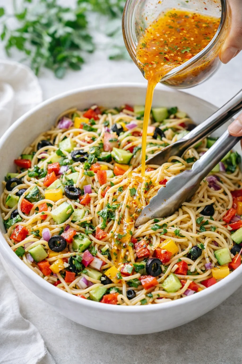Large bowl with cooled spaghetti and chopped vegetables, dressing poured and tongs tossing everything.