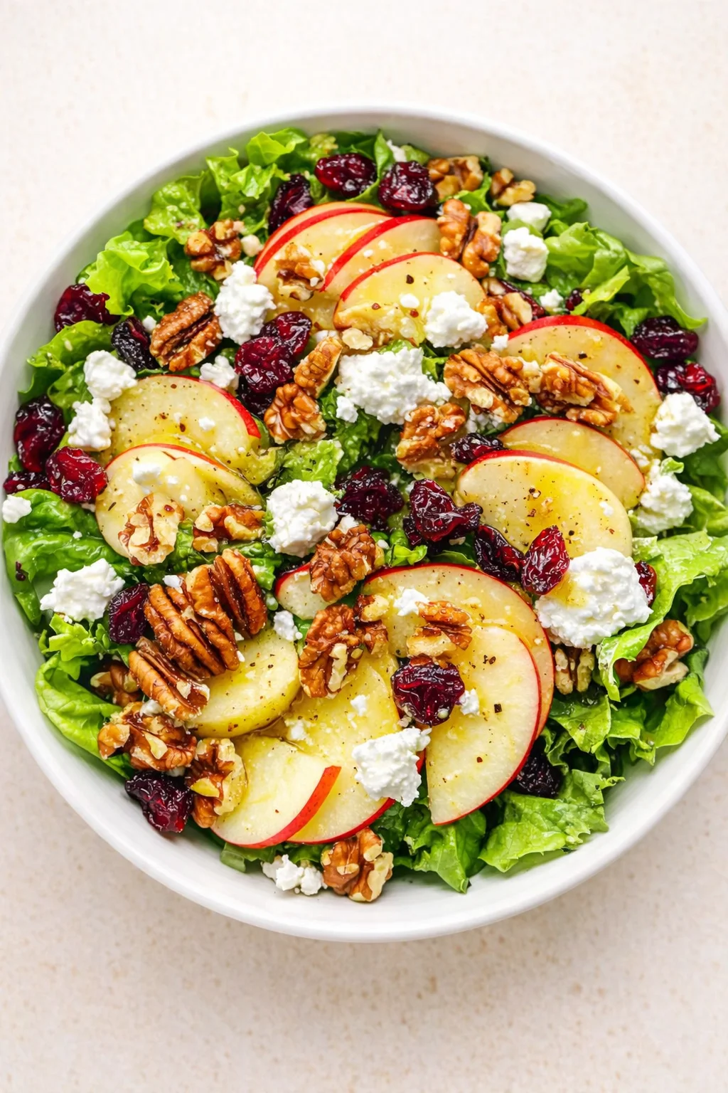 Bright green lettuce base with sliced red apples, dried cranberries, walnuts, and white goat cheese in a bowl.