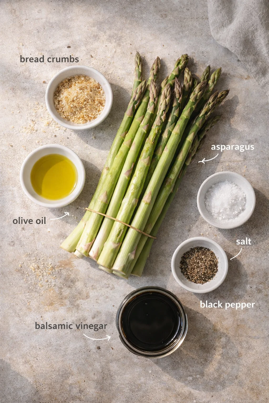 Fresh asparagus bundles with bowls of bread crumbs, olive oil, balsamic vinegar, salt, and pepper on a gray surface.