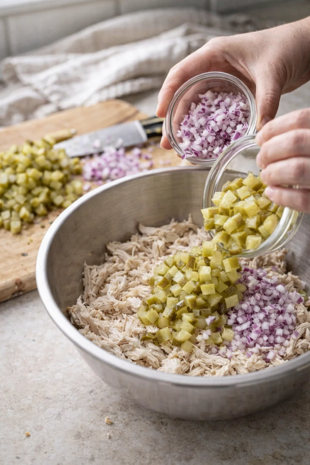 Close-up hands pouring diced pickles and purple onions into shredded chicken in a metal bowl.
