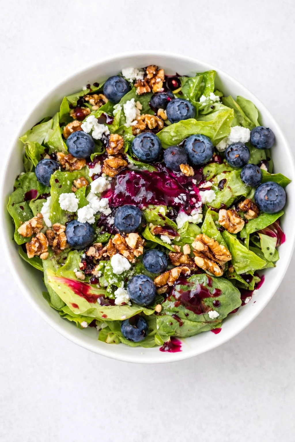 Top-down view of a green salad with blueberries, walnuts, feta, and beet dressing in a white bowl.