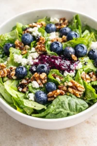 white bowl of mixed greens topped with blueberries, feta, and walnuts