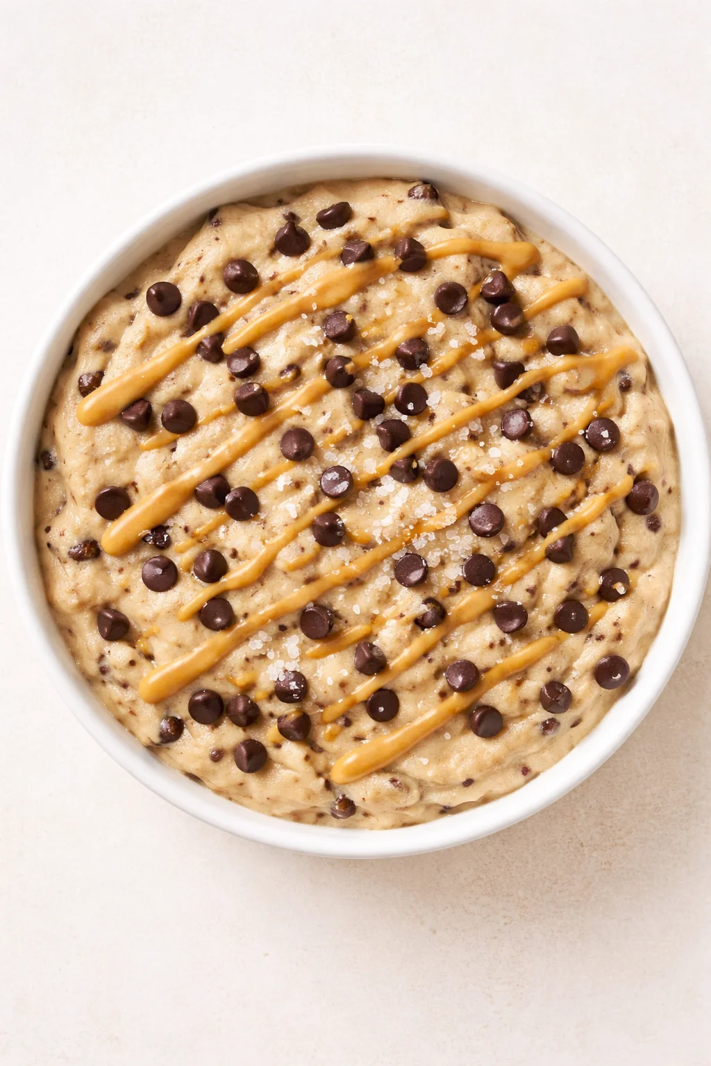 round white bowl of caramel-drizzled cookie dough topped with chocolate chips on a pale textured background