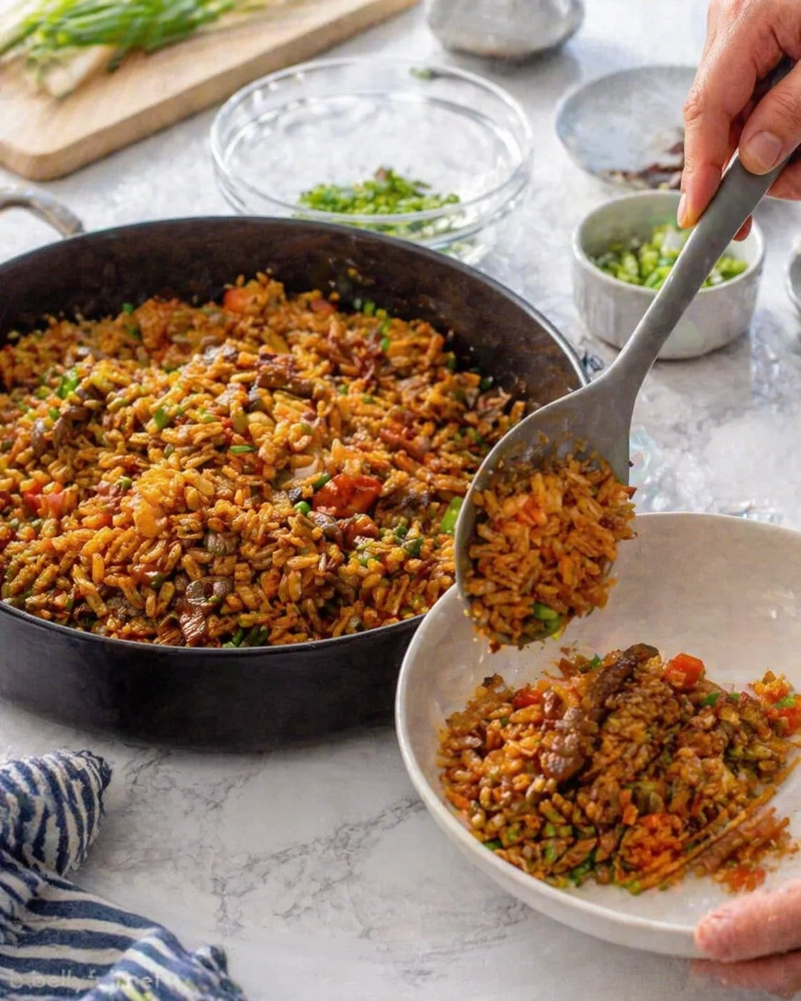 Hand scooping colorful vegetable fried rice from a large skillet into a white bowl.
