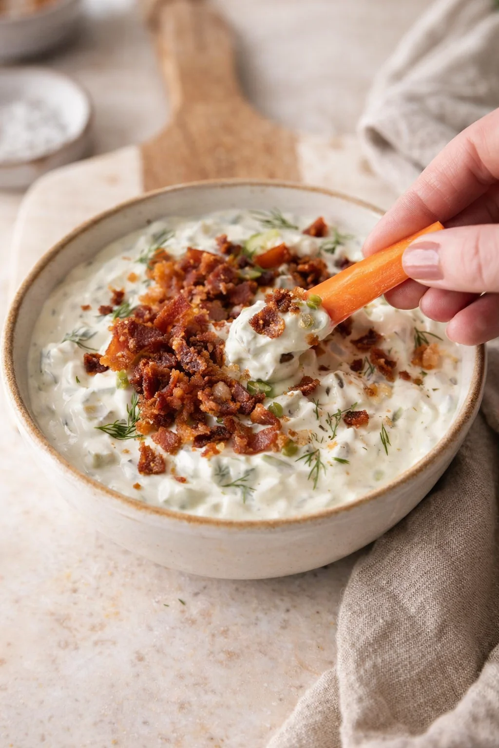Creamy dill-infused dip topped with crumbled bacon, being dipped by a carrot.