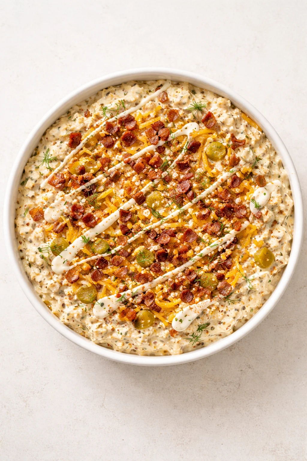 Overhead view of a white bowl filled with creamy, herb-speckled dip topped with bacon, olives, and drizzle.