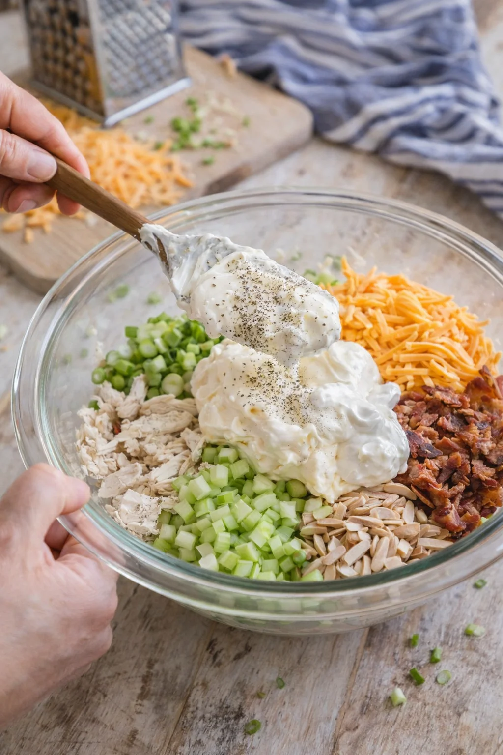 Glass bowl with chopped celery, green onions, chicken, cheese, almonds, and creamy dressing being spooned.