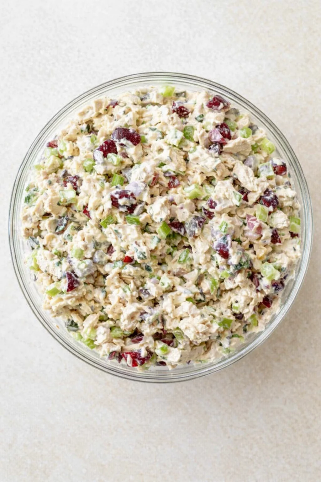 Top-down view of a glass bowl filled with creamy chicken salad, with celery, red grapes, and herbs