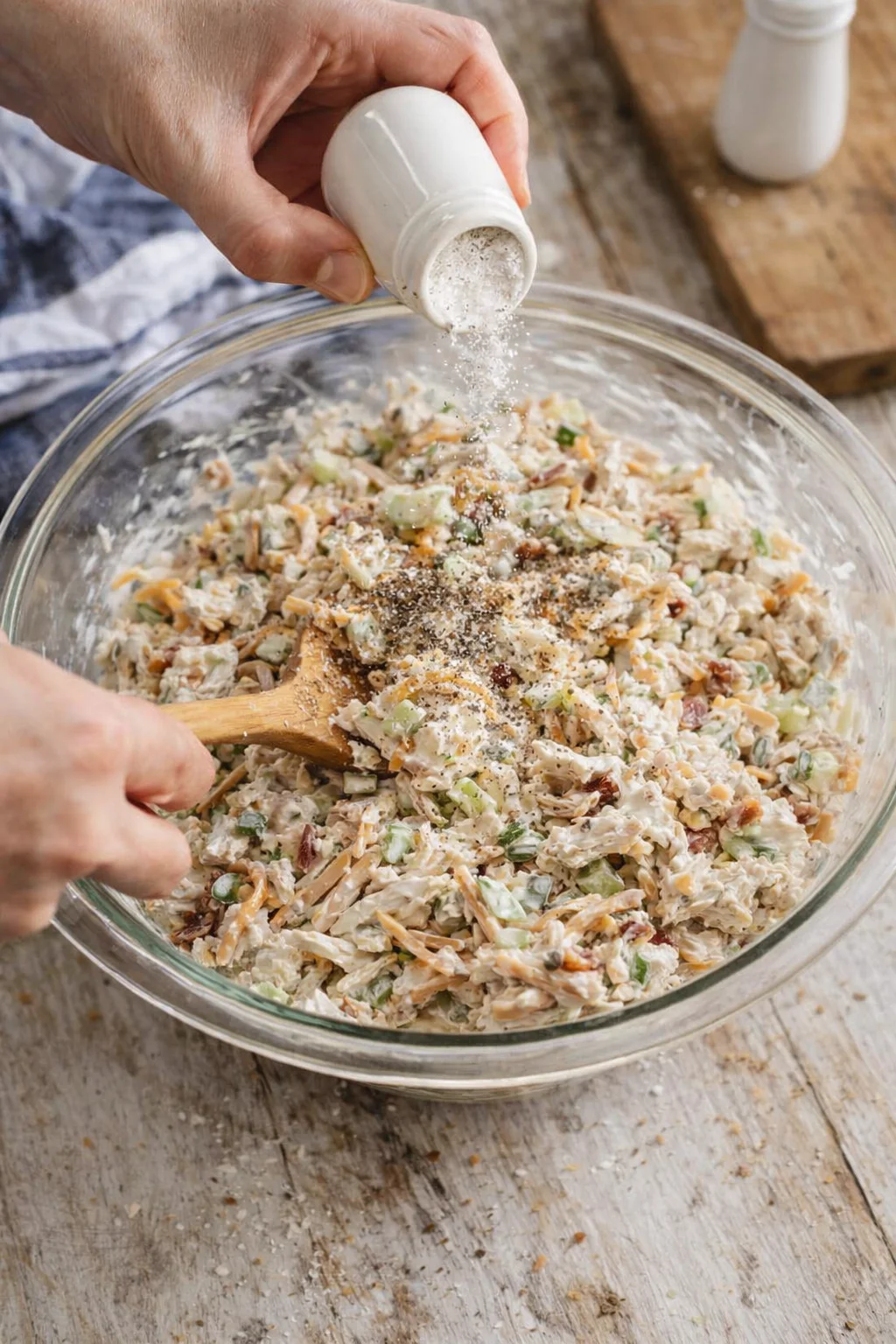 hand sprinkling salt over a glass bowl of creamy chopped salad with celery and onions