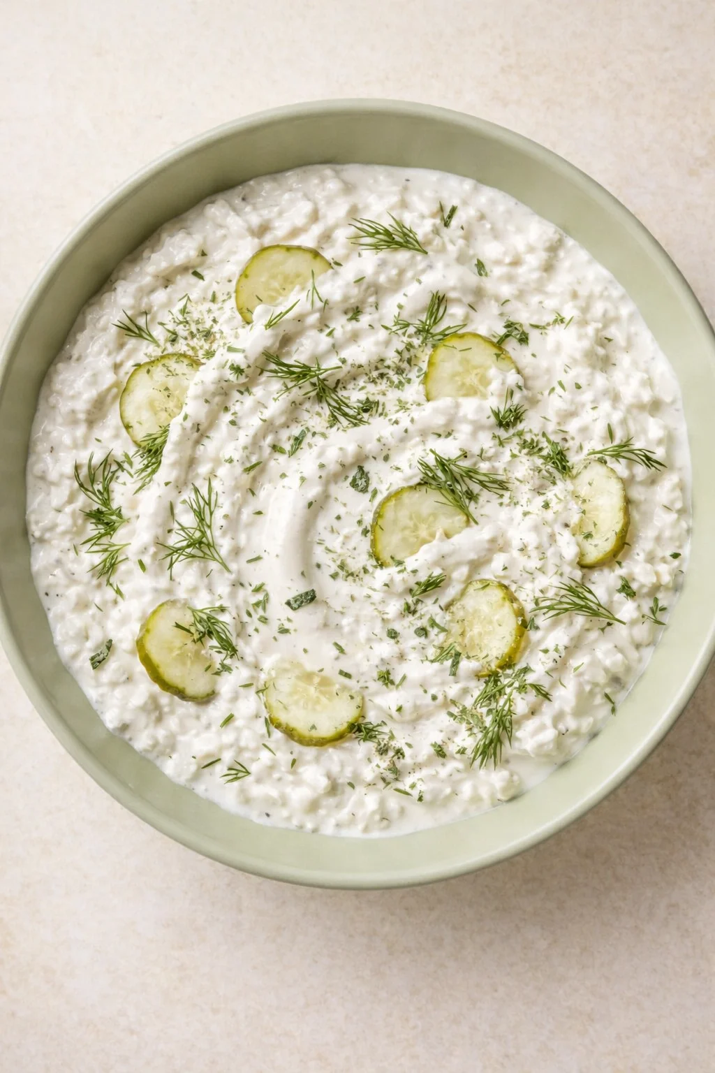 Creamy white yogurt dip with cucumber slices and dill in a pale green bowl.