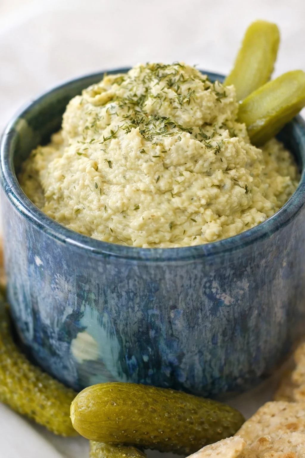 Blue ceramic bowl filled with creamy dill dip, garnished with dill; pickles nearby.
