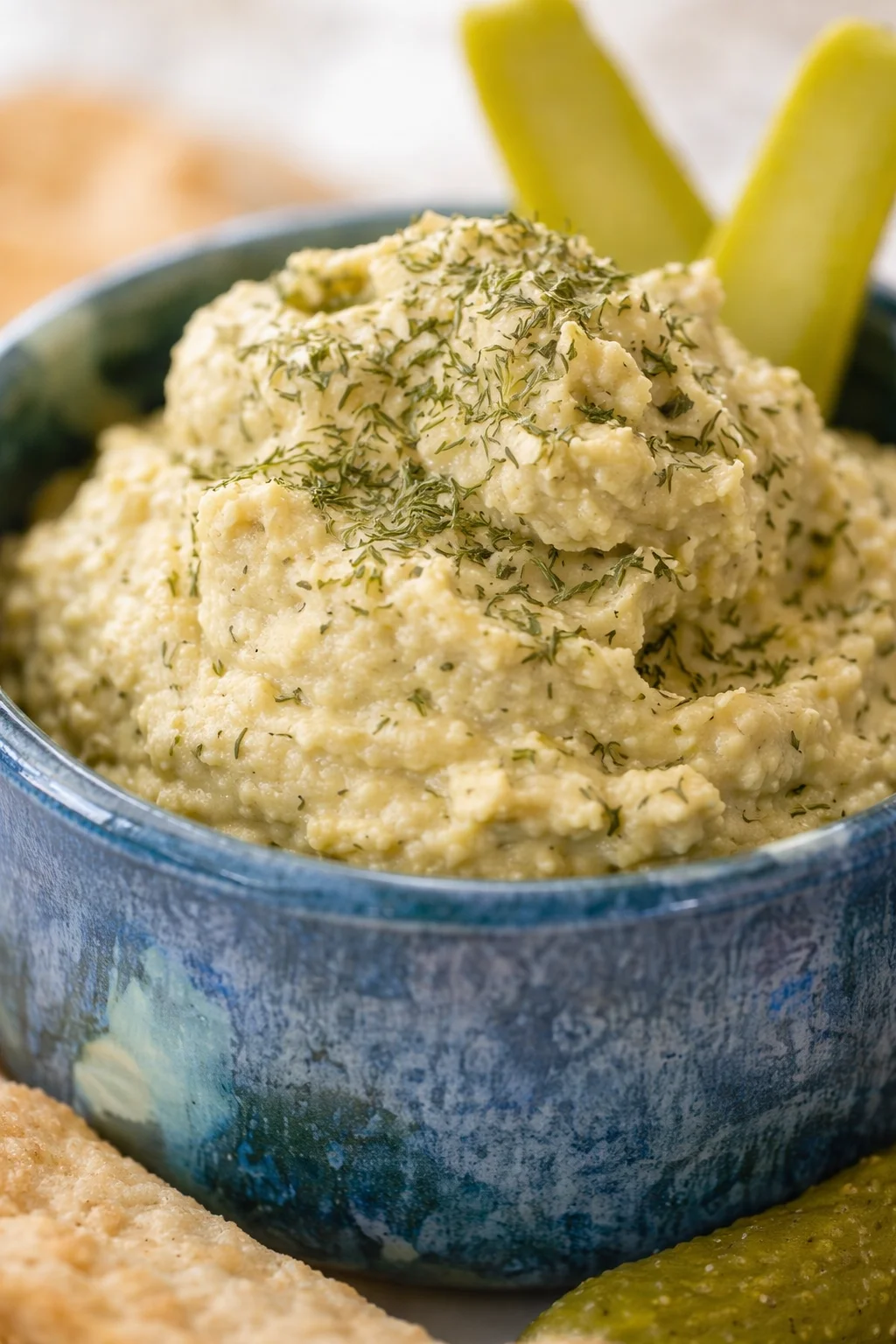 Creamy hummus topped with chopped dill in a blue ceramic bowl with pickle spears in the background.