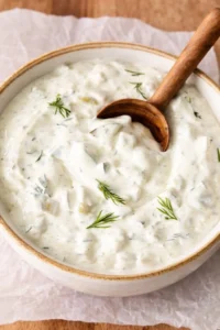 Creamy tzatziki sauce with dill in a white ceramic bowl, wooden spoon resting in the mixture.