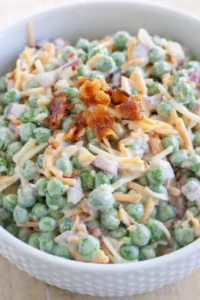 Creamy pea salad with bacon bits and shredded cheese in a white bowl.
