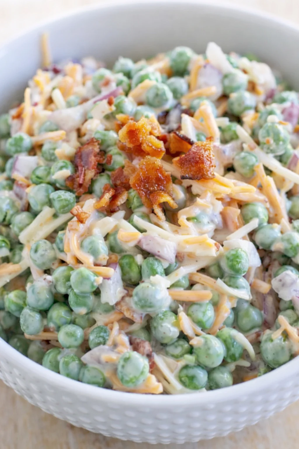 Creamy pea salad with bacon bits and shredded cheese in a white bowl.