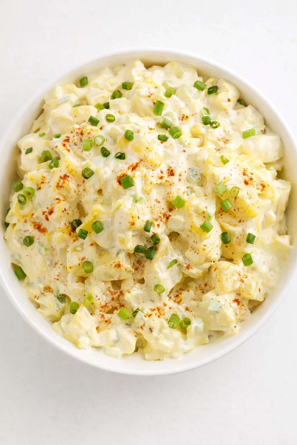white bowl of creamy potato salad topped with chopped green onions and paprika