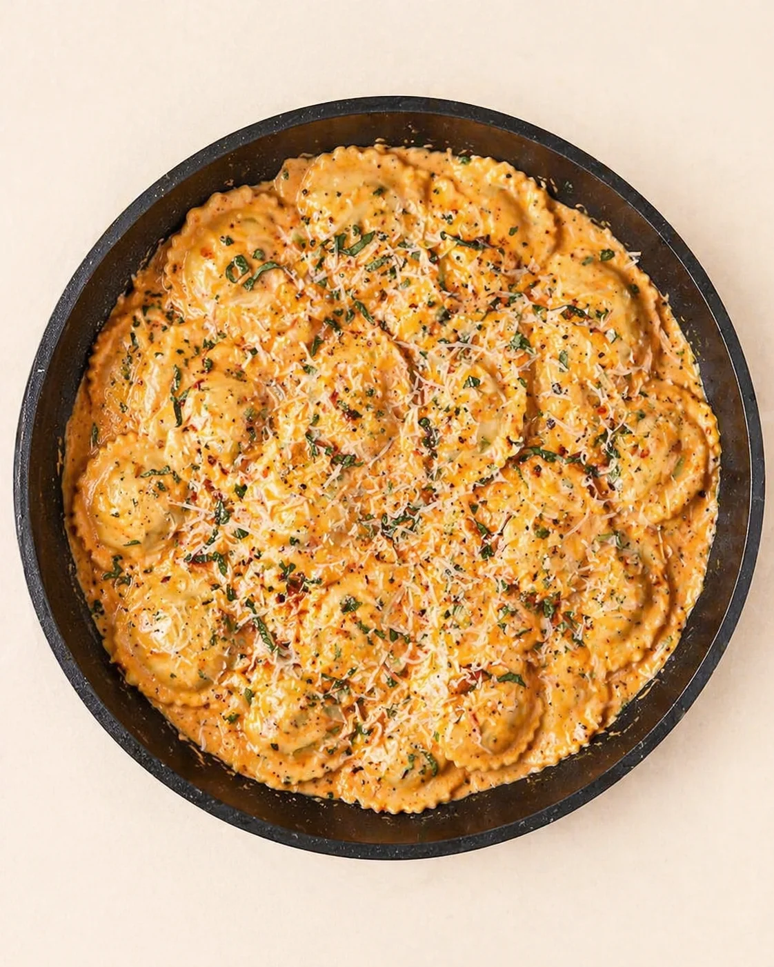 top-down view of a round skillet with ravioli in orange creamy sauce, topped with cheese and herbs